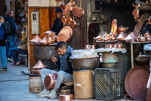 An artisan crafts copperware in a lively Moroccan bazaar in Fez featuring traditional copper stalls and local visitors, capturing the vibrant culture, craftsmanship, and livelihood found in the heart of Morocco.