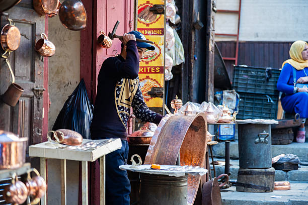 An artisan crafts copperware in a lively Moroccan bazaar in Fez featuring traditional copper stalls and local visitors, capturing the vibrant culture, craftsmanship, and livelihood found in the heart of Morocco.