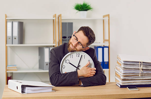 istock Tired, overworked office worker sitting at desk with paperwork, holding clock and sleeping 2222450118