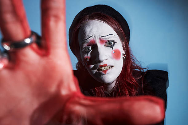 Caucasian young adult woman with red hair wearing mime makeup reaching hand toward camera, expressive face showing dramatic emotion, creative performance concept at home