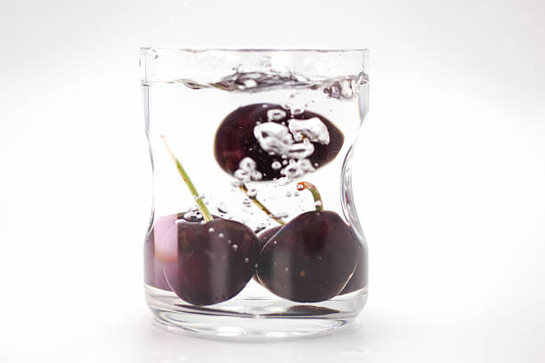 Fresh cherries are dropped into a glass of clear water, creating bubbles and a visually engaging moment as the fruit sinks and interacts with the liquid.