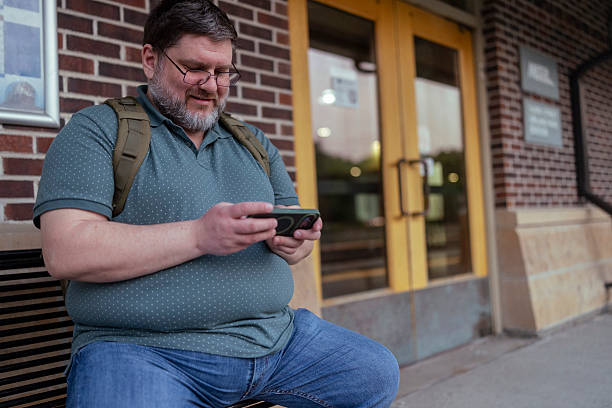 istock Plus size man with a backpack is sitting on a bench and waiting for the train. While waiting for the train, he plays video games on his smartphone 2222457222