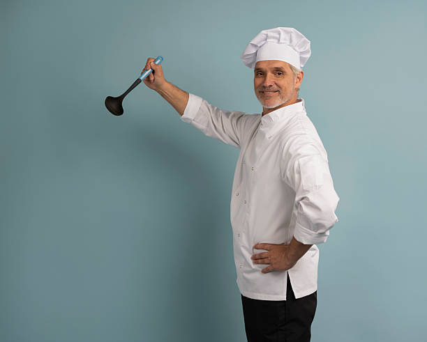 istock Mature male chef with spoon tasting dish on light blue background. 2222466403