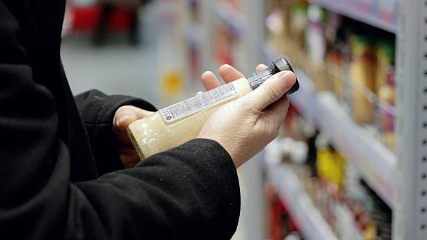 Consumer examining condiment label, comparing nutritional details while selecting product in supermarket aisle, highlighting mindful grocery shopping and health conscious behavior