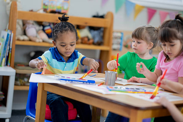 Preschool-aged children are seated around a shared table, enthusiastically participating in creative art activities using bright colors in a cheerful, stimulating learning environment.