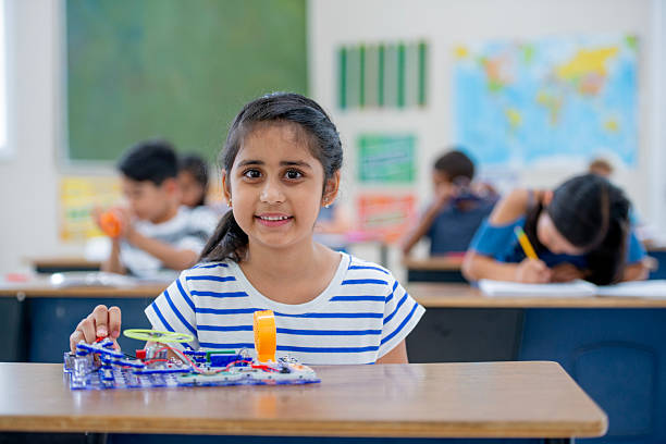 Young girl at her desk in a vibrant classroom engaged in an interactive STEM activity, fostering education and interest in science and technology. Background features children working diligently on their educational tasks.