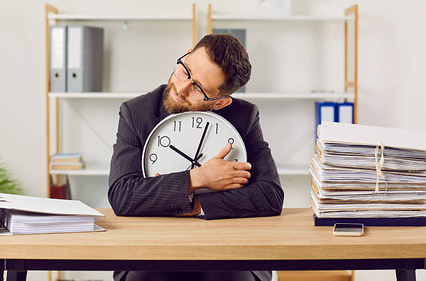 istock Funny, tired, overworked bookkeeper sitting at office desk, holding clock, and sleeping 2222490762