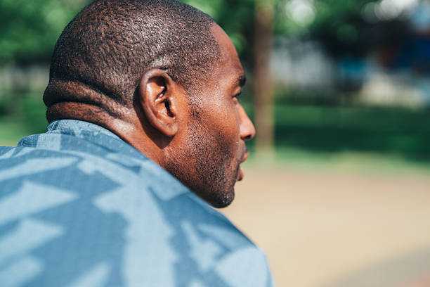 Close-up rear view of a tired man catching his breath after exercising outdoors. Dressed in a patterned athletic jacket, he appears to be recovering from an intense workout in a sunny park. His skin glistens with sweat, and the background is filled with greenery and natural light. The image conveys themes of perseverance, physical effort, recovery, and personal fitness goals. Perfect for illustrating real moments in athletic training, resilience, or wellness journeys.