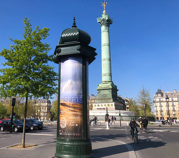 Paris, France - April 10, 2025: The Place de la Bastille, with the Colonne de juillet monument in the background and a Morris column in the foreground; it advertises an exhibition called “Deserts” in the National Museum of Natural History.