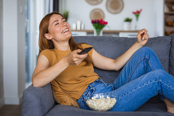istock Young woman laughing while watching tv and eating popcorn on sofa 2222526894