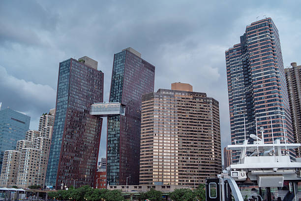 Modern skyscrapers and residential buildings line the waterfront in New York City. The skyline features distinctive glass office towers and luxury apartment buildings with a mix of architectural styles. In the foreground, part of a boat is visible on the water. The scene is captured under a cloudy, overcast sky creating a moody atmosphere. The urban landscape shows the density of Manhattan's waterfront development with buildings of varying heights and designs clustered together.
