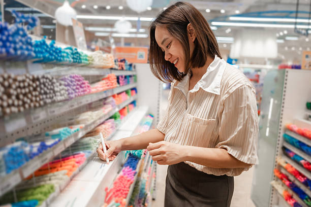 Smiling Asian saleswoman jotting down notes on her notepad while surrounded by a vibrant array of stationery in a bustling store