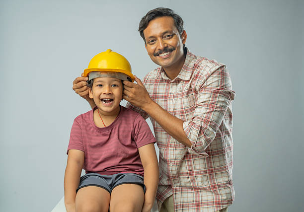 Portrait of smiling mid adult engineer father wearing hardhat to his excited son while standing against white background