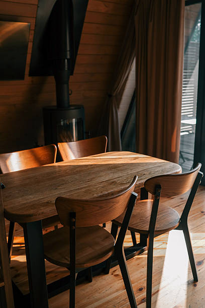 istock Wooden dining table with matching chairs in a cozy interior, illuminated by warm sunlight. A modern black wood stove stands in the background beside a large window with beige curtains 2222556065