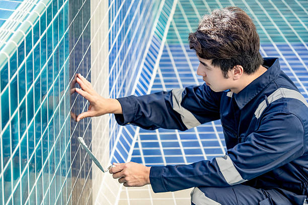 Asian man construction worker using trowel or building spatula spreading cement grout on blue ceramic tiles in the swimming pool