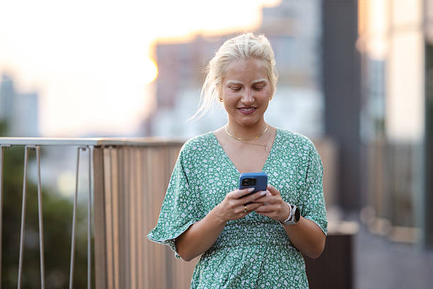 istock Smiling young woman using smartphone while standing on a sunny balcony 2222572555