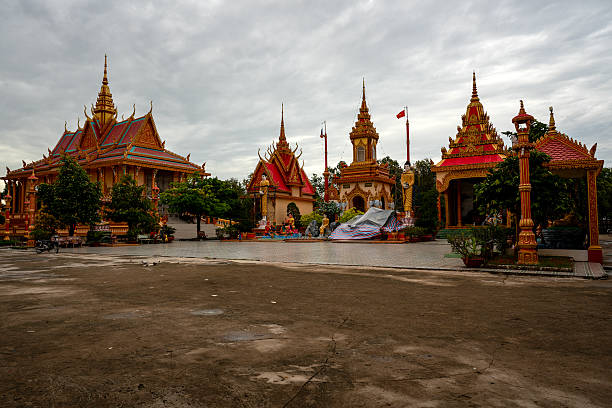 Bac Lieu, Vietnam – August 26, 2019: A wide view of a traditional Buddhist temple complex featuring ornate golden rooftops, statues, and a central staircase. The temple grounds are empty under a cloudy sky, with trees and religious structures arranged across the open courtyard.
