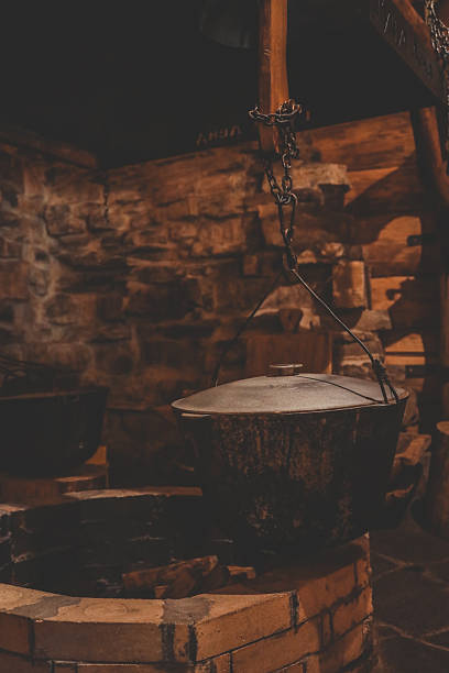 istock Iron cauldron with lid hangs above stone fire pit inside rustic log-and-stone cabin, evoking traditional hearth cooking amid warm, shadowy ambient light 2222581927