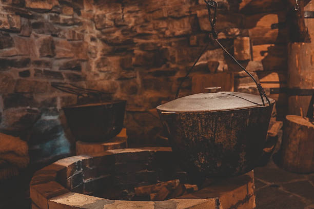 istock Iron cauldron with lid hangs above stone fire pit inside rustic log-and-stone cabin, evoking traditional hearth cooking amid warm, shadowy ambient light 2222581942
