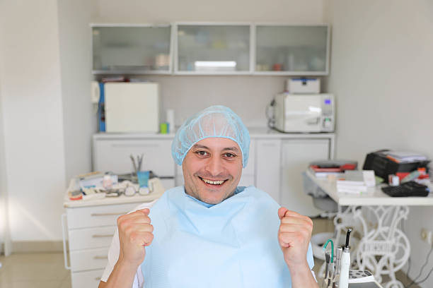 A happy male patient in a dental chair wearing a protective cap and bib, raising his fists in celebration after a successful dental procedure. The scene conveys satisfaction, relief, and trust in professional healthcare
