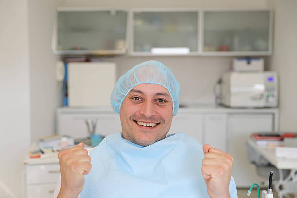 A happy male patient in a dental chair wearing a protective cap and bib, raising his fists in celebration after a successful dental procedure. The scene conveys satisfaction, relief, and trust in professional healthcare