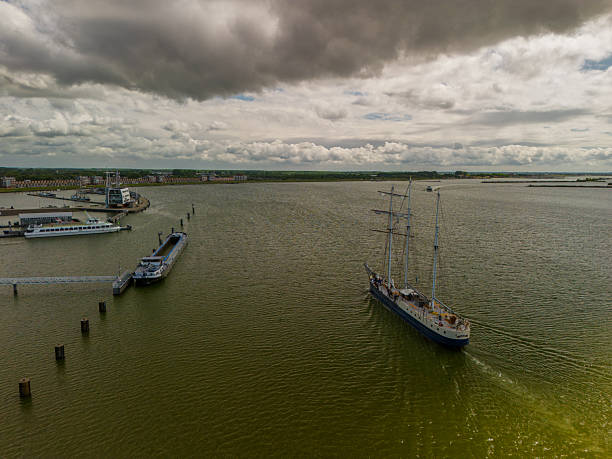 A majestic sailing ship glides across calm waters under a partly cloudy sky, with coastal landscapes and a modern dam structure visible in the background.