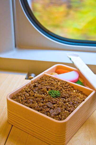 An overhead shot of a makunouchi bento featuring chicken rice (torimeshi) as the main component. The bento box is a rectangular plastic container with multiple compartments. The chicken rice is a mix of rice and small pieces of chicken, with a slightly orange hue. The bento also includes a variety of side dishes, such as fried vegetables, a boiled egg cut in half, pickled ginger, and a dark-colored vegetable. The bento is placed on a dark surface.