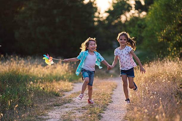 Rear view of two young girls with their arms spread straight out to each side pretending to be airplanes as they walk down a rural gravel driveway. Focus is on the younger sister in the blue denim dress with curly red hair. Her left hand is holding small flowers she picked along the driveway. Her older sister is on the left side of the image with a blue and white paisley dress and curly blonde hair. Both girls are wearing cowboy boots.