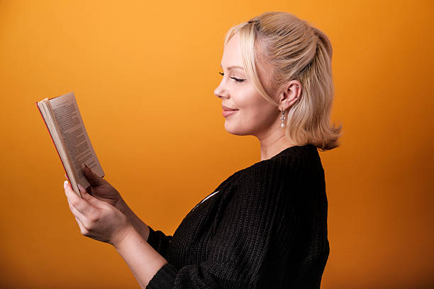 Side view portrait of a blond woman enjoying reading a book, smiling serenely with an orange background, creating a peaceful and engaging scene