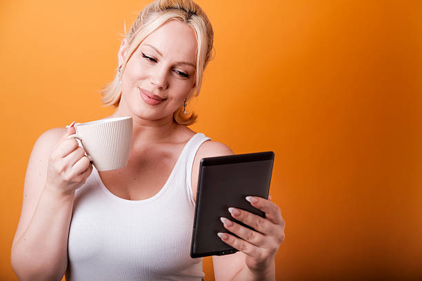 Happy woman savoring morning coffee while reading an e-book on a tablet, surrounded by a vibrant orange background, embodying the essence of modern lifestyle and technology