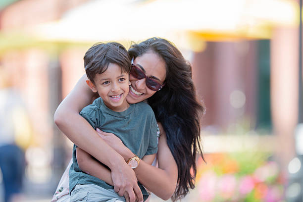 A candid moment capturing a mother happily embracing her young son in an outdoor setting, expressing love and tenderness. Vibrant background blurred with sunlight creates a warm, cheerful atmosphere.