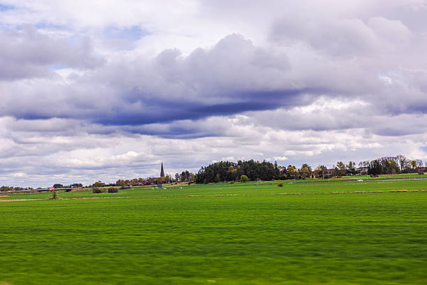 Green spring fields with distant church steeple under dramatic cloudy sky in rural Sweden.