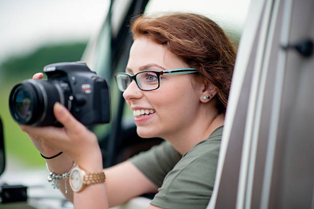 A smiling woman with glasses holds a camera while leaning out of a vehicle window, capturing scenic moments during a road trip. Her excitement and passion for photography are apparent.