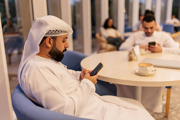 Muslim man in kandura and his friend using smart phones while sitting at a chic Dubai café.