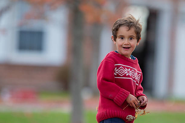Young boy wearing a red sweater smiling joyfully outside amidst vibrant fall foliage.