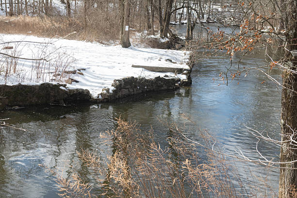 This is a photograph of a snow covered landscape reflected in the water’s surface of a woodland stream in Ramapo Reserve on a winter day in Mahwah, New Jersey.