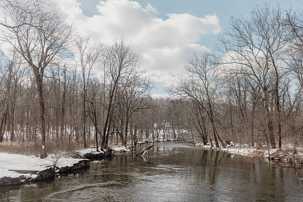 This is a photograph of a snow covered landscape reflected in the water’s surface of a woodland stream in Ramapo Reserve on a winter day in Mahwah, New Jersey.