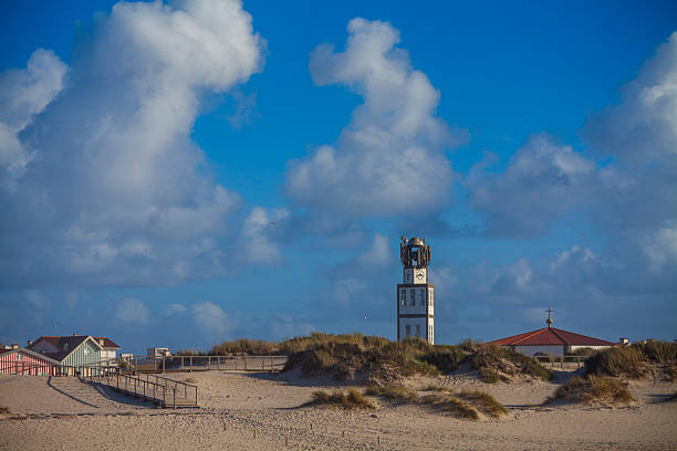 A tall, white clock tower with a distinct top stands prominently amidst sandy dunes and scattered buildings under a bright blue sky filled with dramatic white clouds, showcasing a unique coastal landscape