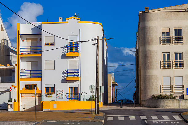 A brightly painted white and yellow modern building with blue railings stands prominently on a street corner, contrasting with a more subdued neighboring building under a clear blue sky, capturing urban architectural diversity