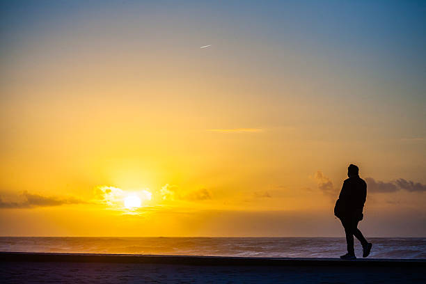 istock A lone figure walks along a seaside path at sunset, silhouetted against a vibrant orange sky 2222651569