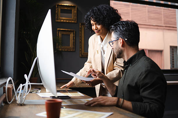 Two designers collaborating on a project, reviewing documents and discussing ideas in a modern office environment Creative professionals analyzing project details, sharing insights while working together in contemporary workspace with laptop and paperwork website design stock pictures, royalty-free photos  images