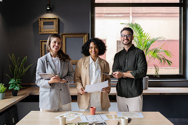 istock Three young architects are smiling at the camera while holding plans and a digital tablet in their modern office 2222660222
