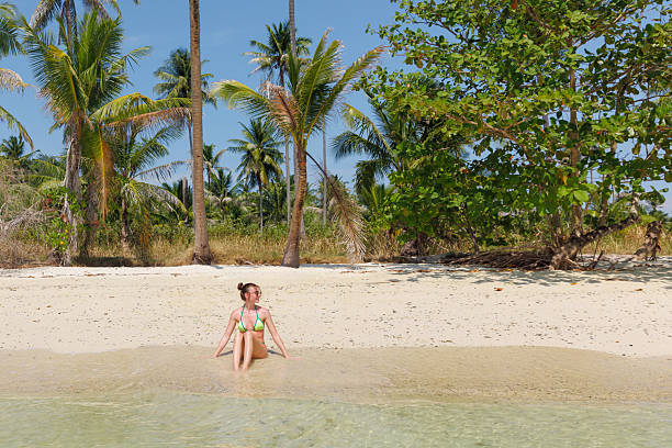 A woman enjoys a sunny day at a beautiful beach in Thailand, sitting in shallow waters with soft sand and swaying palm trees surrounding her. Bright beach towels are laid out nearby.