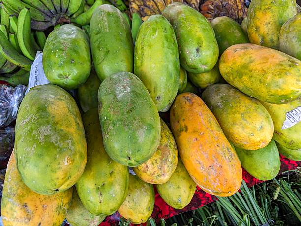 The image shows fresh tropical fruits – pineapple and papaya. In the center, there is a green bowl filled with sliced papaya pieces, while in the background, a halved papaya with black seeds and a whole pineapple are visible. The vibrant fruit colors create an appetizing and natural look