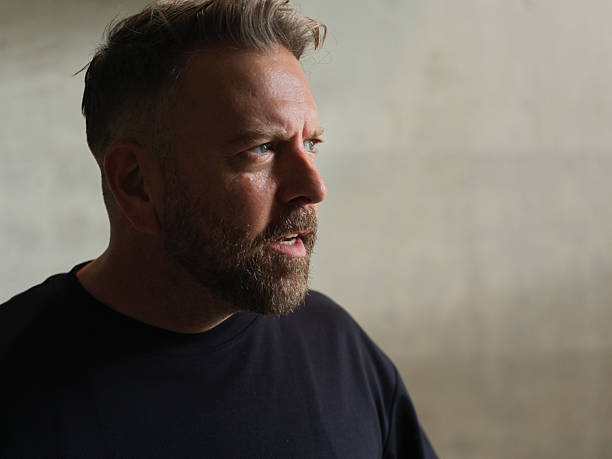 Side profile portrait of a mature man with a trimmed beard and short hair, wearing a dark t-shirt and standing against a plain textured wall. The man appears to be in deep thought or mid-conversation, with natural light casting soft shadows on his face, adding intensity to the scene. Ideal for themes such as emotion, contemplation, decision-making, and masculinity.