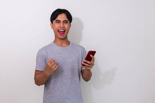 istock Excited young Asian man celebrating success while shouting and using a mobile phone, isolated on white background 2222688538