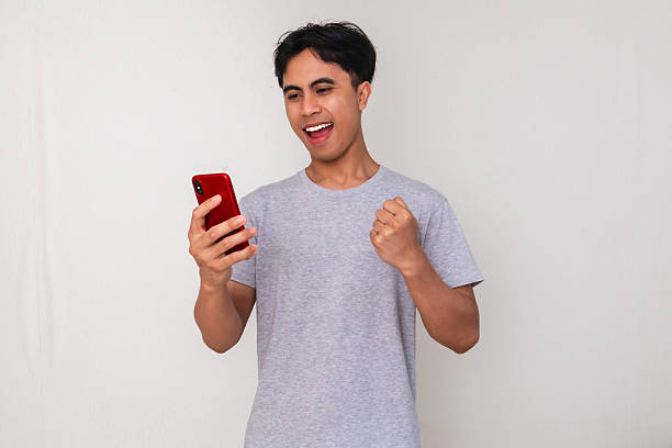 istock Excited young Asian man celebrating success while shouting and using a mobile phone, isolated on white background 2222688558