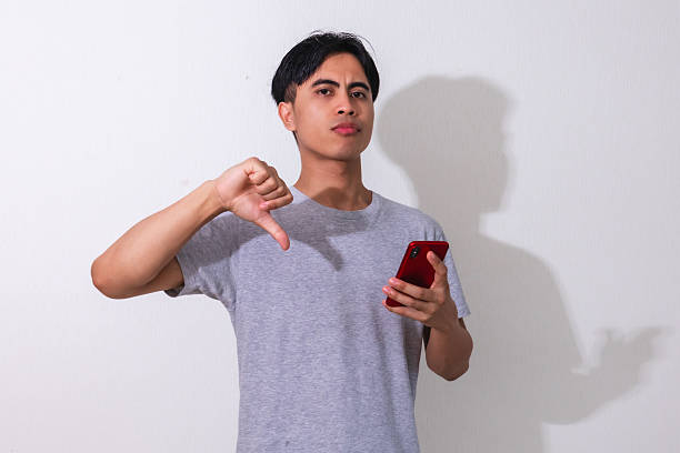 istock Disappointed young Asian man showing thumbs down while holding a smartphone, isolated on white background, perfect for advertising, technology, and reaction concepts 2222688572