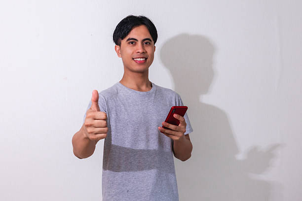 istock Smiling young Asian man showing thumbs up while holding a smartphone, isolated on white background. Perfect for promotion, mobile ads, or tech-related campaigns 2222688614