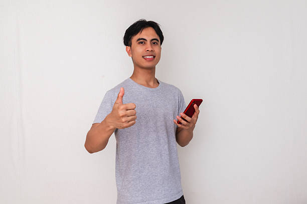 istock Smiling young Asian man showing thumbs up while holding a smartphone, isolated on white background. Perfect for promotion, mobile ads, or tech-related campaigns 2222688673